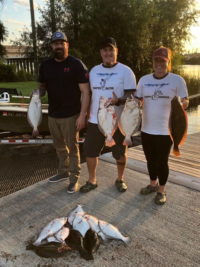 Three people proudly display their caught fish while standing on a wooden dock by the water.
