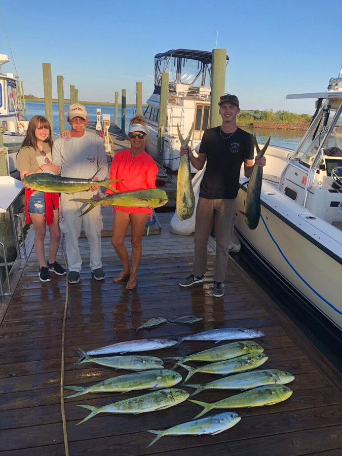 Three people proudly display their caught fish while standing on a wooden dock by the water.
